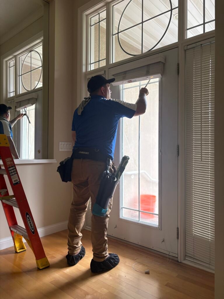 man in blue shirt next to a ladder cleaning the interior side of windows and glass door