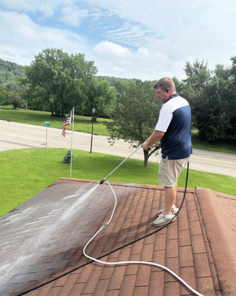 man standing on a roof wearing tan pants blue and white shirt, spraying the roof to clean it