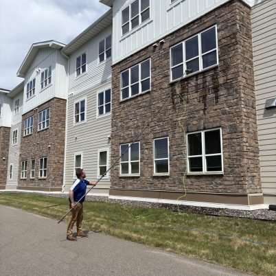 man standing in a street or drive holding a long pole to clean 2nd story windows of a 3 story building
