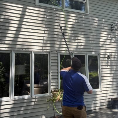 man with blue shirt and tan pants facing a tan house washing the 2nd story windows with a long pole