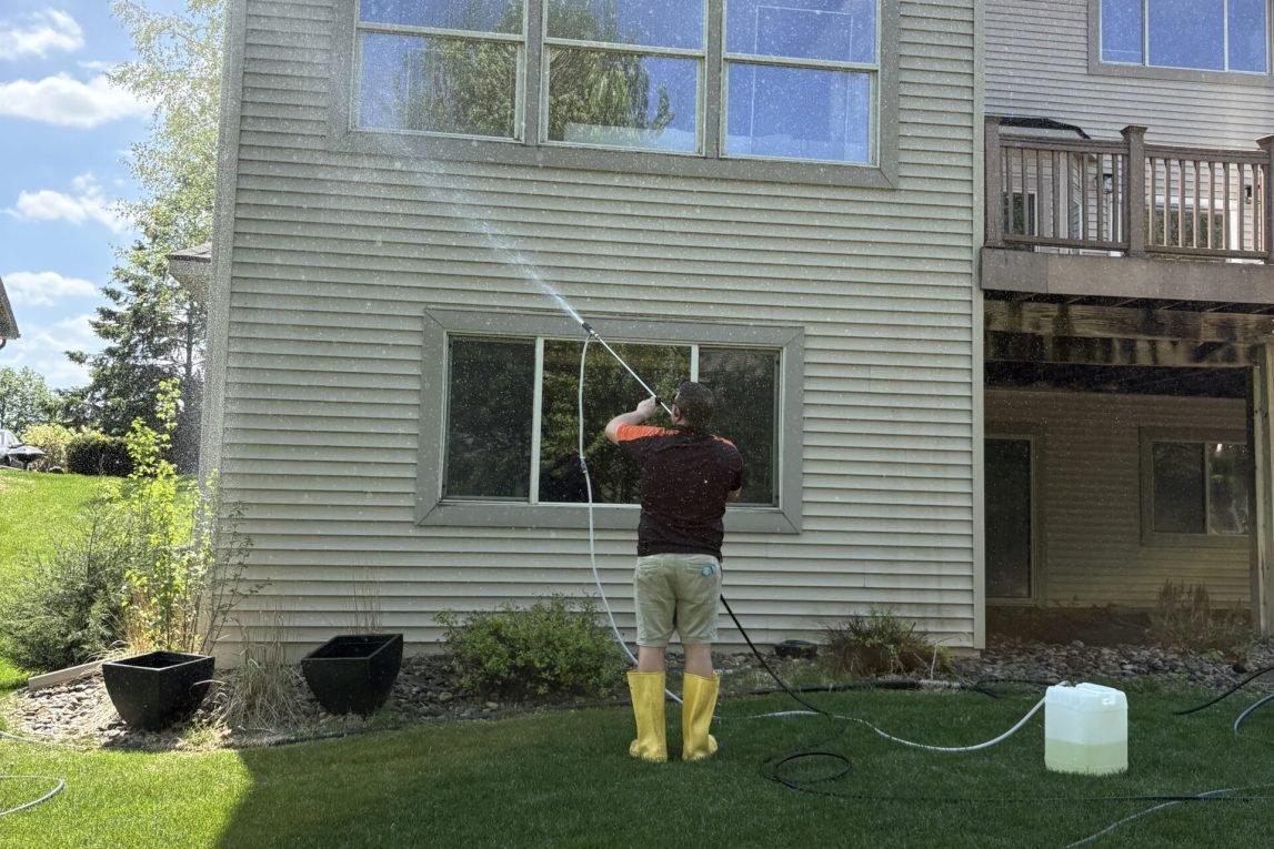 Man standing in the grass next to a house, spraying the siding to soft wash