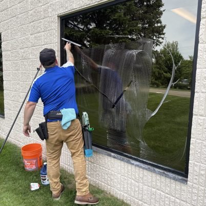 man in blue shirt, black toolbelt, tan pans, next to an orange bucket cleaning the exterior side of a window