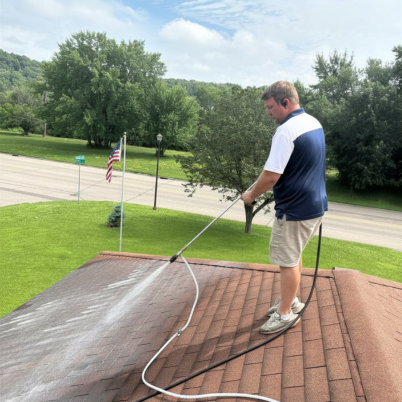 man standing on a roof wearing tan pants blue and white shirt, spraying the roof to clean it