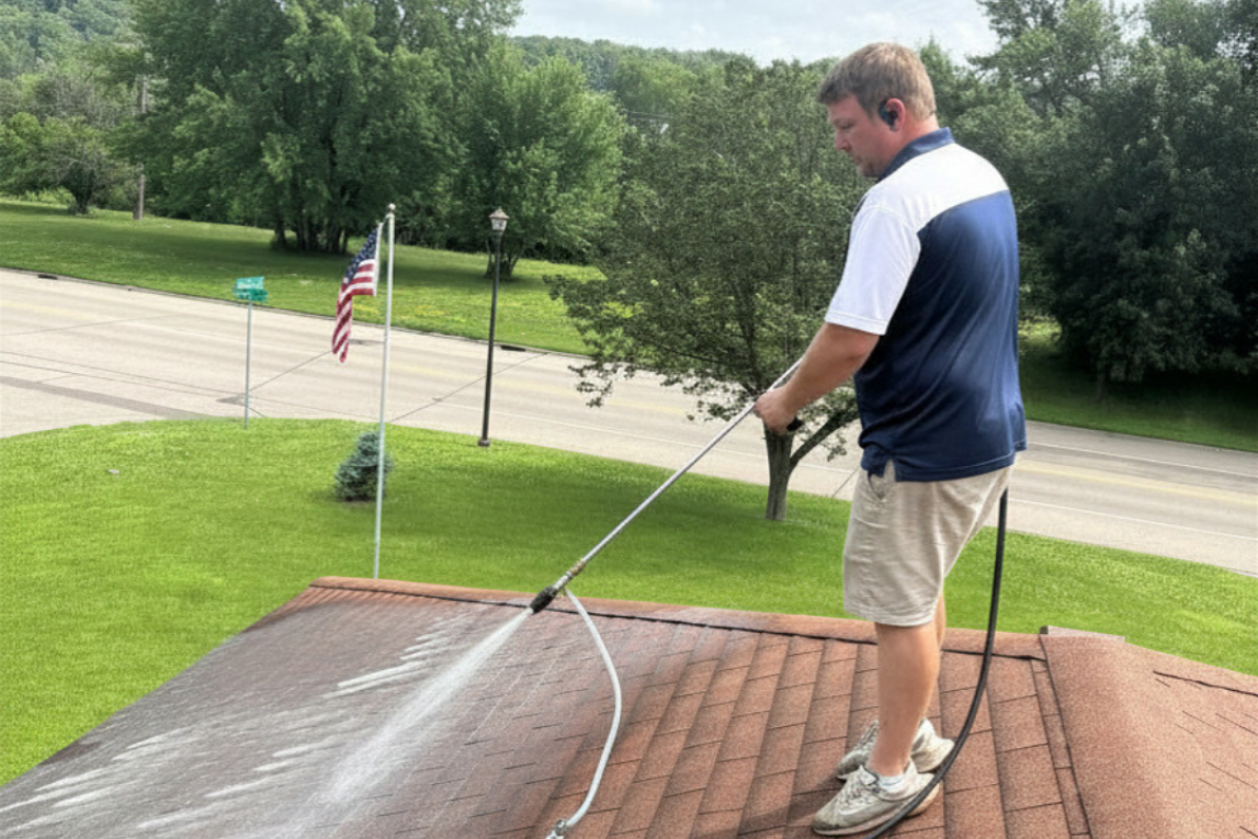 man standing on a roof wearing tan pants blue and white shirt, spraying the roof to clean it