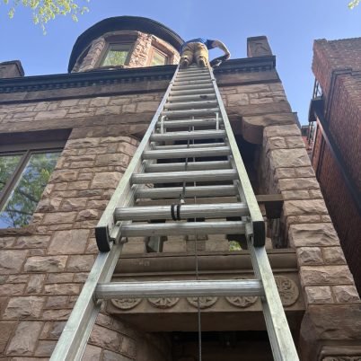 tall brick home, large ladder leaded against the brick, man at the top of the ladder cleaning gutters