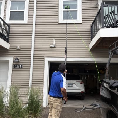man wearing a blue shirt and tan pants holding a long pole to clean windows on a second story home
