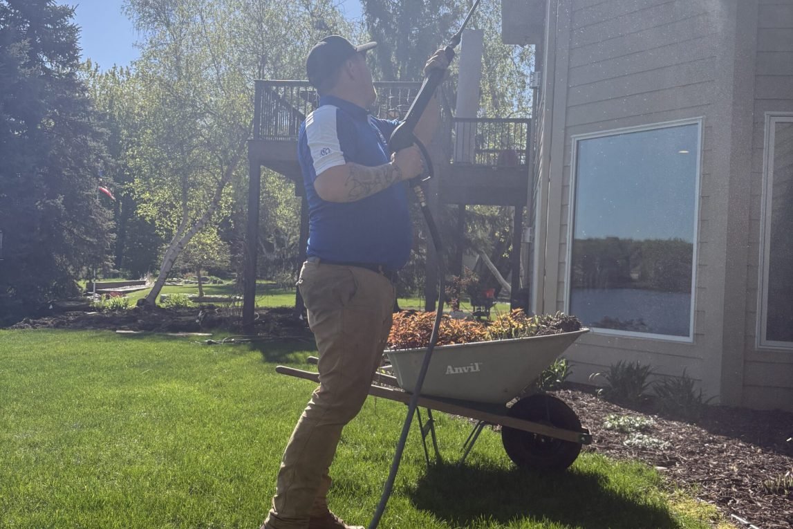 man wearing blue shirt, holding a hose, spraying the side of a house