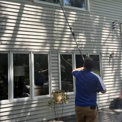 man in blue shirt and tan pants facing a home holding a long pole looking up to clean windows on the second floor of a home