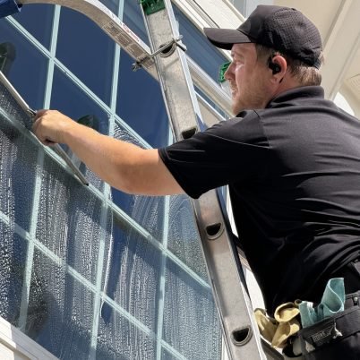 man in a black shirt and black hat, standing on a tall ladder cleaning the exterior side of windows