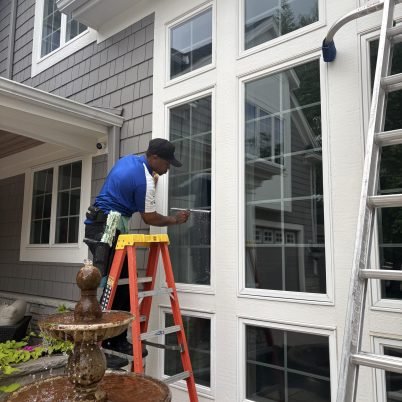 man in blue shirt, black pants and hat, standing on an orange ladder with a yellow top next to large windows cleaning them