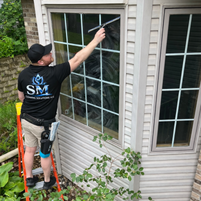 man on ladder wearing a black shirt with white and blue logo that reads SM Cleaning Solutions, he is using a squeegy on the window