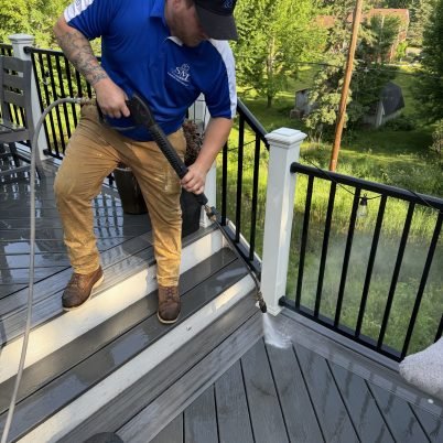 man in blue shirt, black hat, tan pants, spraying a grey and white deck surface