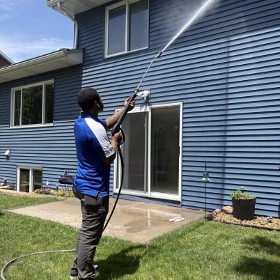 man in blue shirt, grey pants, spraying the side of a blue two story home