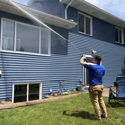 man in blue shirt, tan pants, black hat, spraying a blue house